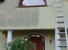 Fasada domu jednorodzinnego w&nbsp;trakcie czyszczenia. Widoczny mocny kontrast: lewa strona ściany nad&nbsp;wejściem jest brudna i&nbsp;pokryta zielono-szarym nalotem, podczas gdy&nbsp;prawa strona jest już umyta i&nbsp;ma&nbsp;jasnobeżowy kolor. O&nbsp;czystą część ściany oparta jest wysoka aluminiowa drabina.