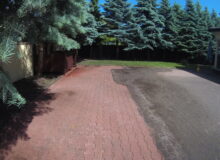 Podjazd z&nbsp;kostki brukowej w&nbsp;trakcie czyszczenia. Widoczny ostry kontrast pomiędzy już umytą, czerwoną kostką po&nbsp;lewej stronie, a&nbsp;mocno zabrudzoną, szaro-czarną powierzchnią po&nbsp;prawej. W&nbsp;tle zadbany ogród z&nbsp;rzędem wysokich drzew iglastych.