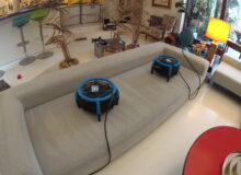 Długa, jasnoszara sofa materiałowa po&nbsp;profesjonalnym czyszczeniu. Zabrudzenia zniknęły, a&nbsp;na&nbsp;czystym siedzisku stoją dwie niebieskie dmuchawy przyspieszające suszenie tkaniny. W&nbsp;tle przestronny salon.